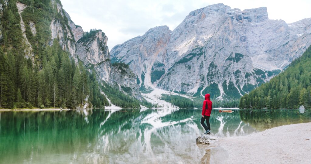 Photo of man stood on shore of a lake surrounded by mountains