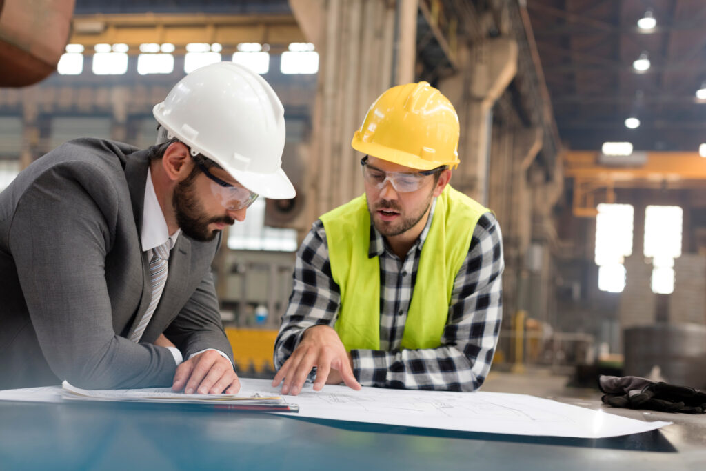 Two men wearing hard hats reviewing documents in a factory setting