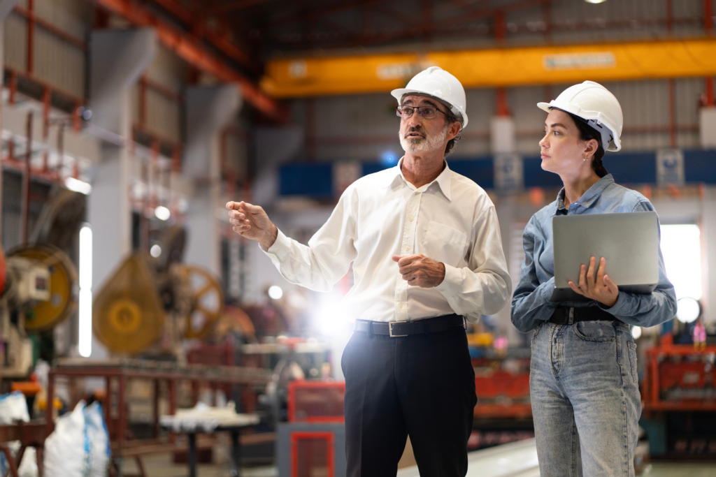 Two people in a factory, wearing helmets. Machinery is visible in the background.