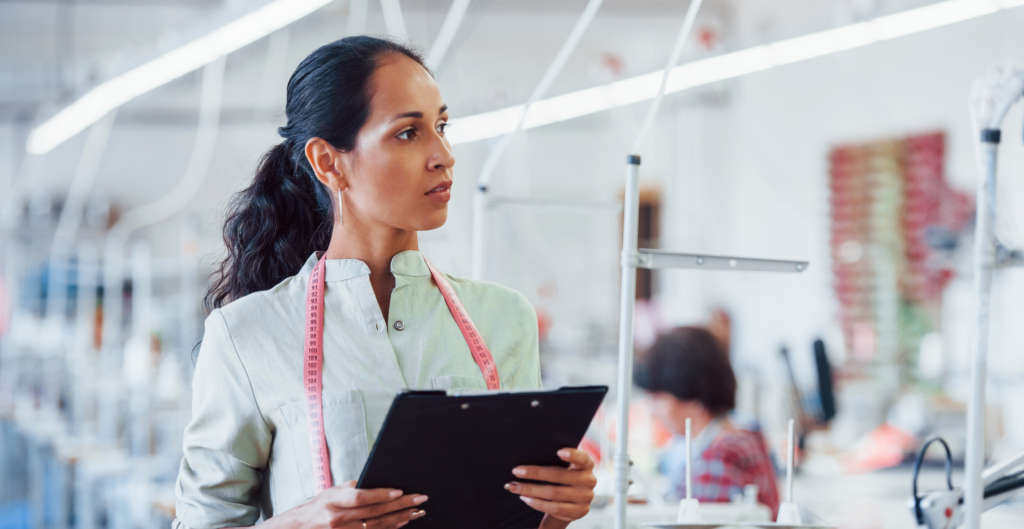 A woman in a white coat checks equipment while holding a clipboard.